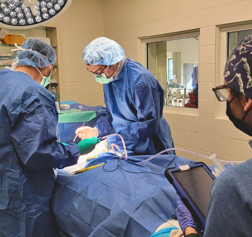 Three medical professionals in surgical attire are shown in an operating room, focused on performing surgery on a patient. Two are directly involved in the procedure, while the third is observing and holding a tablet. The environment is sterile with medical equipment visible.