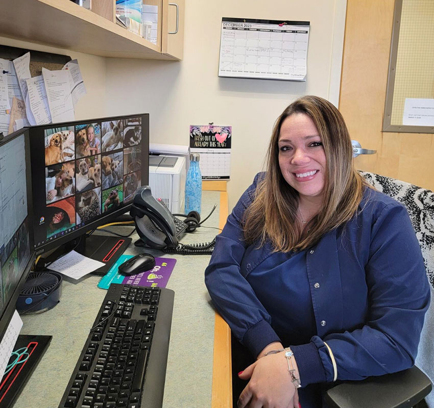 A woman with long hair sits smiling at a desk in an office. She is wearing a dark blue jacket and is surrounded by office equipment, including a dual-monitor setup displaying various dog pictures, a keyboard, a phone, and a calendar on the wall behind her.