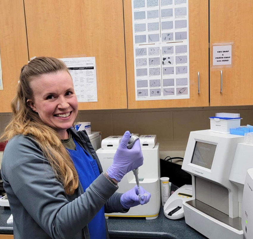 A woman in laboratory attire, including purple gloves and a blue apron, smiles while using a pipette at a lab bench. Behind her, there are scientific instruments, charts on the wall, and wooden cabinets. The environment indicates it's a research laboratory.