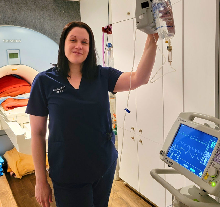 A healthcare professional in navy scrubs stands beside medical equipment in a hospital room. She is holding an IV fluid bag connected to a drip and is standing next to a monitor displaying vital signs. An MRI machine is visible in the background.