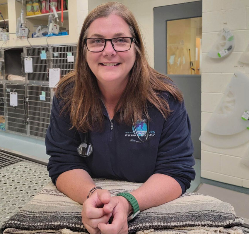 A woman wearing glasses and a navy blue veterinary clinic jacket stands smiling in an animal care facility. She has shoulder-length brown hair and rests her hands on a padded table. Behind her are various medical supplies and equipment.
