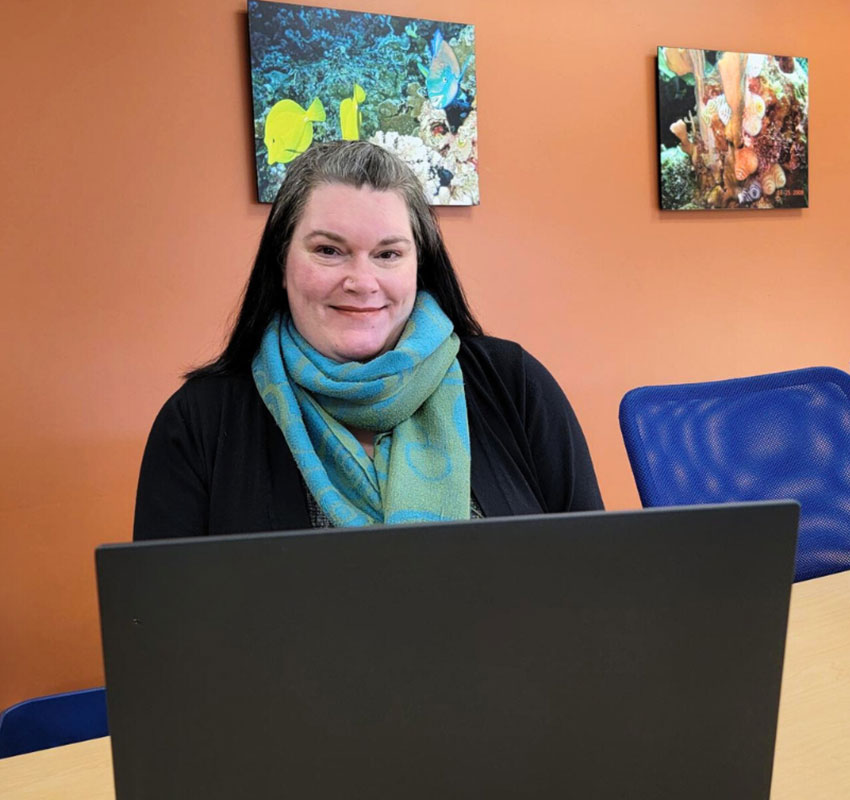A person with long dark hair sits at a table with an open laptop. They are smiling and wearing a teal scarf. The background features an orange wall with two framed pictures of underwater scenes.