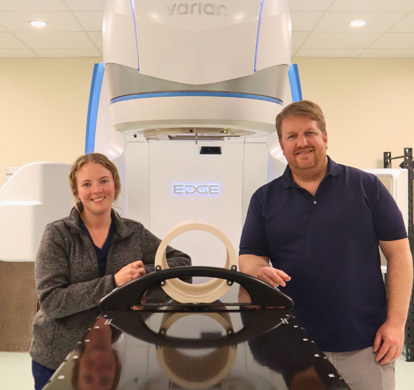 Two people smiling and standing beside a Varian Edge radiation therapy machine in a clinical setting. One person is wearing scrubs, and the other is in casual attire. The background features a brightly lit room with a tiled ceiling.