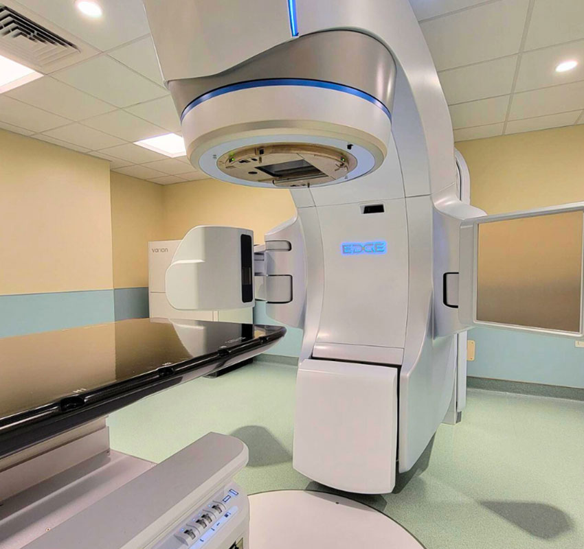 A sleek, modern radiation therapy machine with a large circular structure and an adjustable patient platform in a well-lit medical room with pale green flooring.
