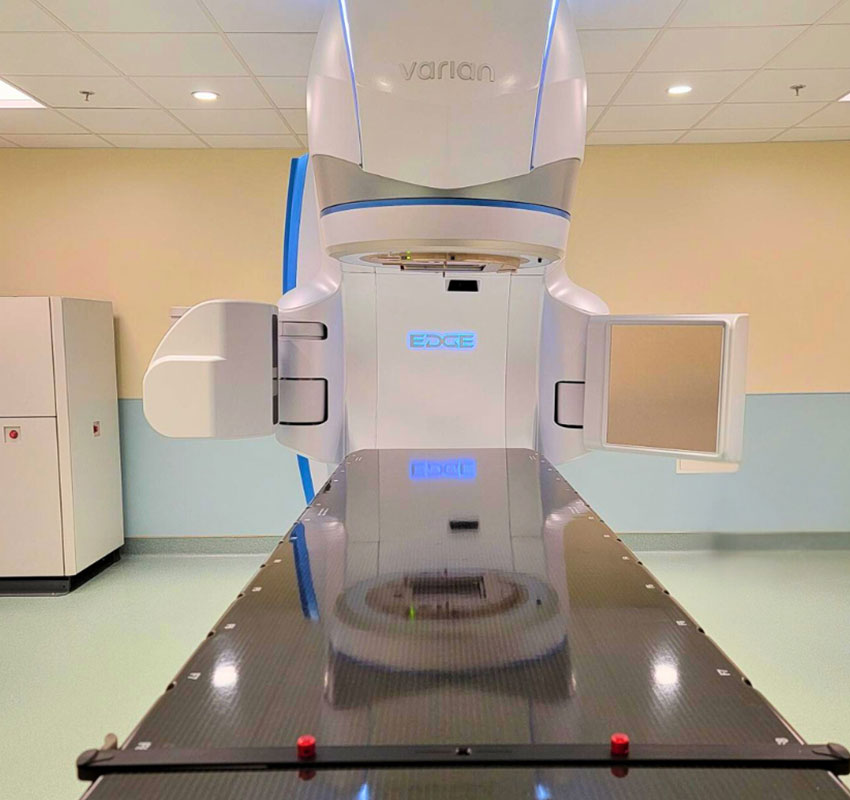 A medical radiotherapy machine with the brand name "Varian" and model "EDGE" is positioned over a treatment table. The room has light-colored walls and a tiled ceiling, suggesting a clinical environment.