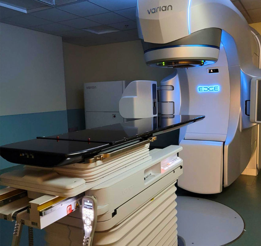 A modern radiation therapy machine, labeled "Varian" and "Edge," with a sleek, futuristic design. It features a large, curved gantry and a horizontal patient treatment table, set in a clinical room.