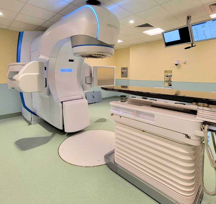 A modern medical room featuring a large, white linear accelerator machine used for radiation therapy. The room has a light green floor and cream walls, with monitors and equipment visible in the background.